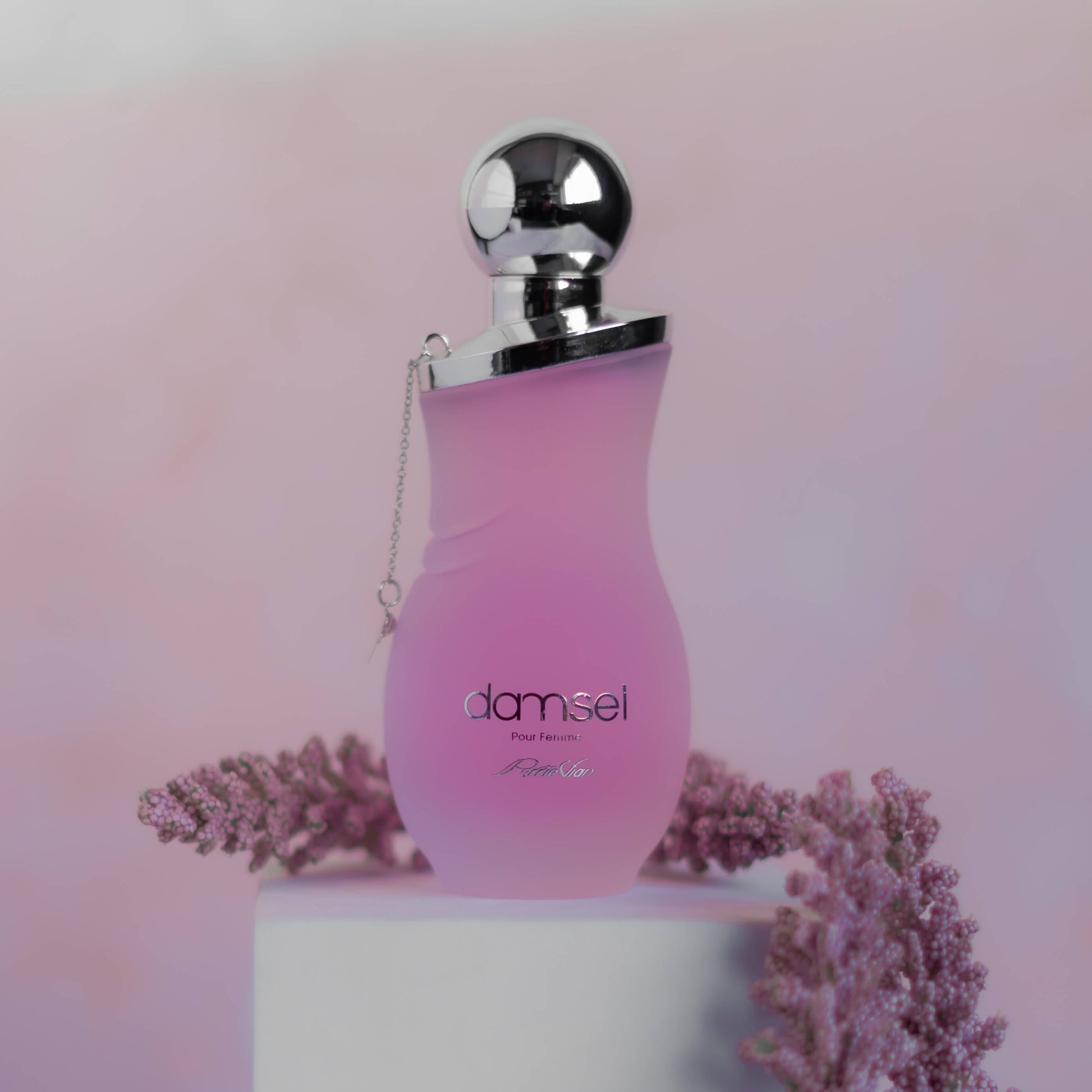 A pink bottle of DAMSEL EDP for women, with a silver cap, displayed on a white surface with purple flowers around it.