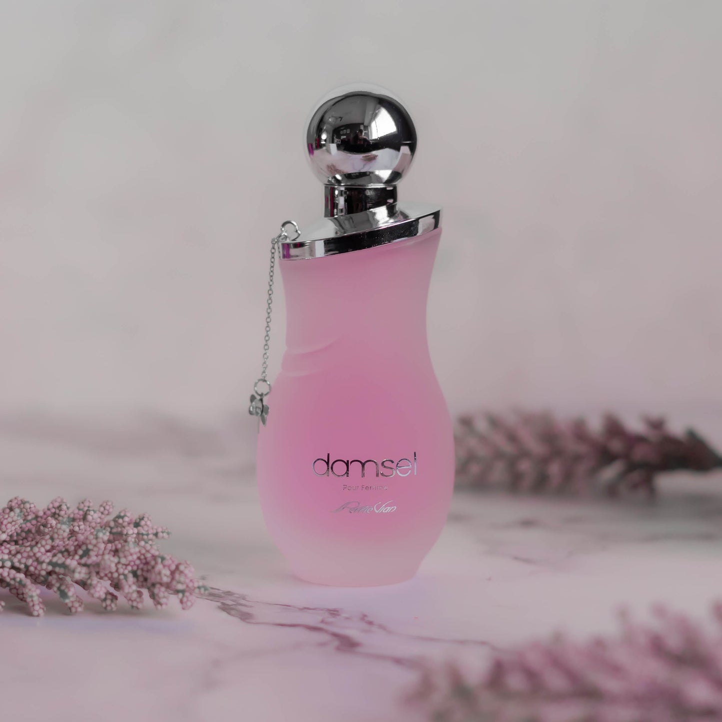 A pink bottle of DAMSEL EDP for women, with a silver cap, displayed on a white surface with purple flowers around it.