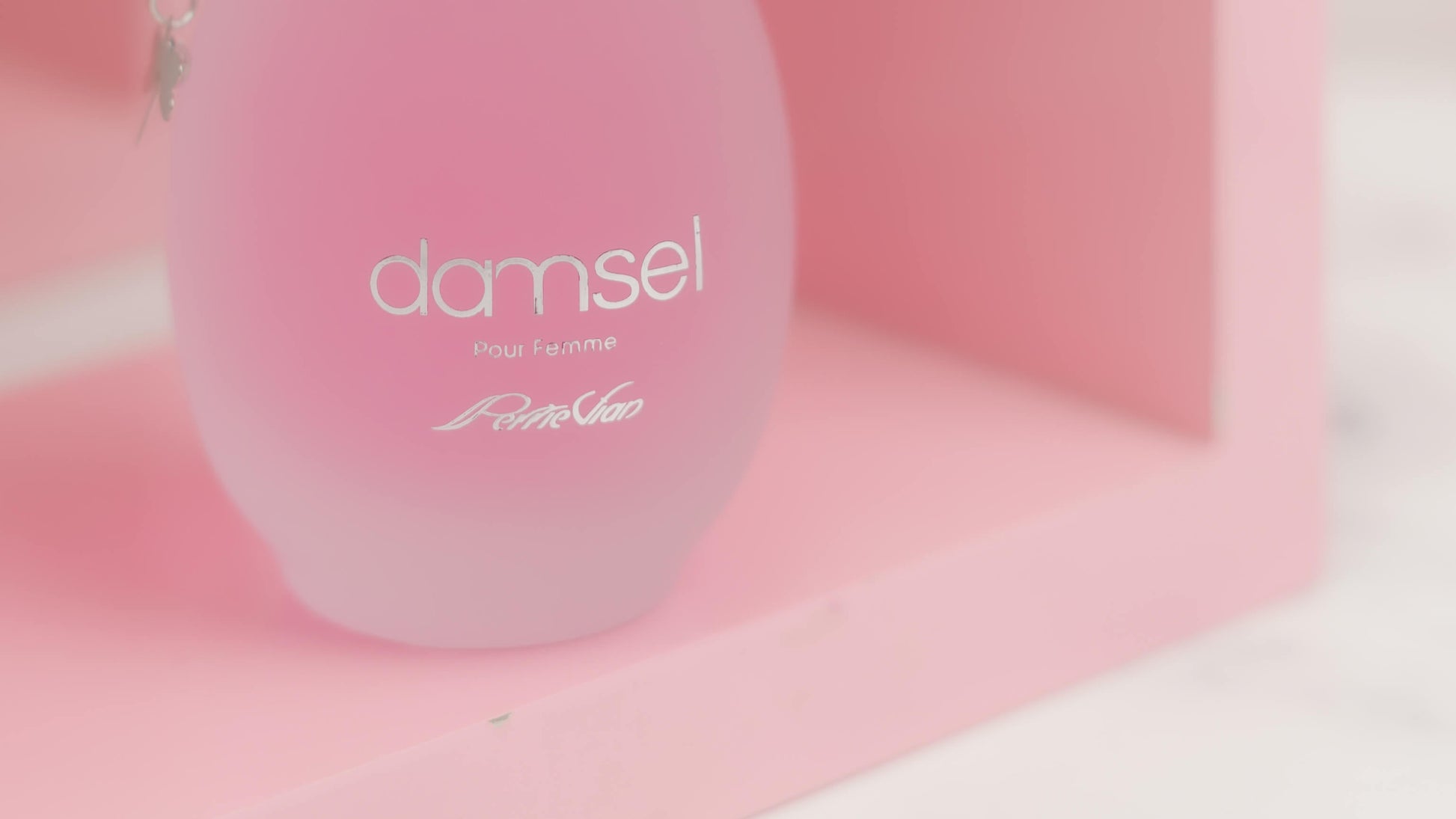 A pink bottle of DAMSEL EDP for women, with a silver cap, displayed on a white surface with purple flowers around it.