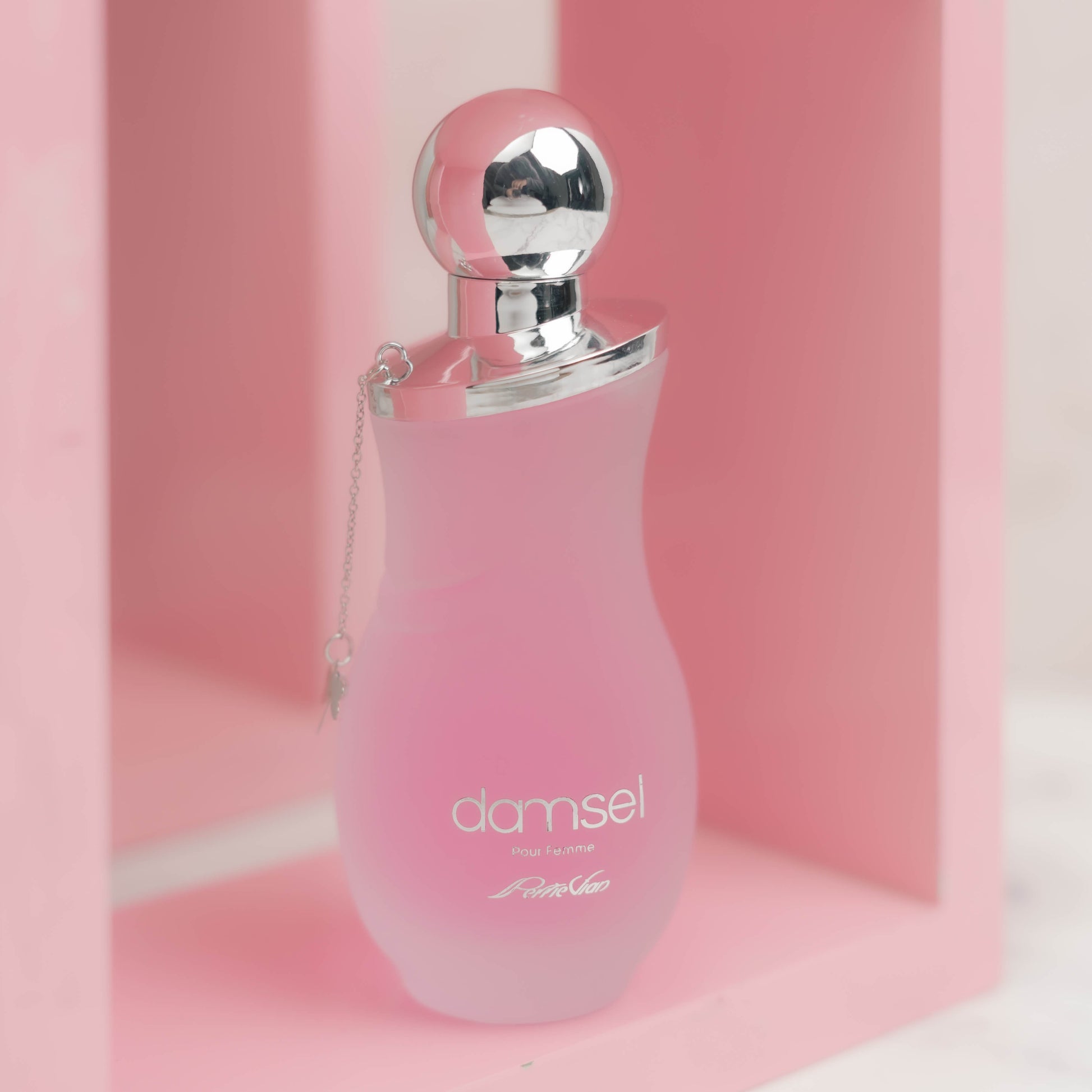 A pink bottle of DAMSEL EDP for women, with a silver cap, displayed on a white surface with purple flowers around it.