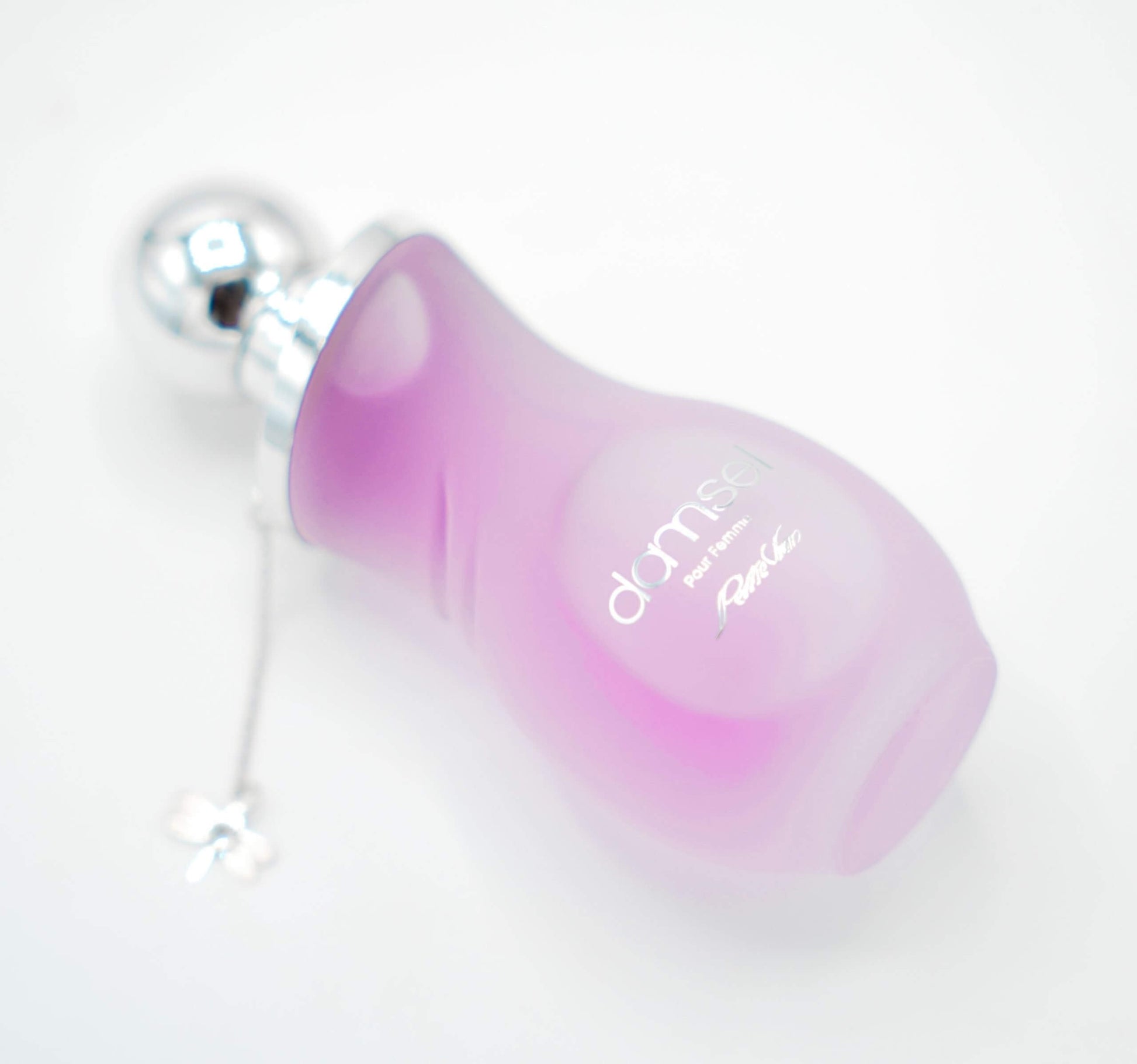 A pink bottle of DAMSEL EDP for women, with a silver cap, displayed on a white surface with purple flowers around it.