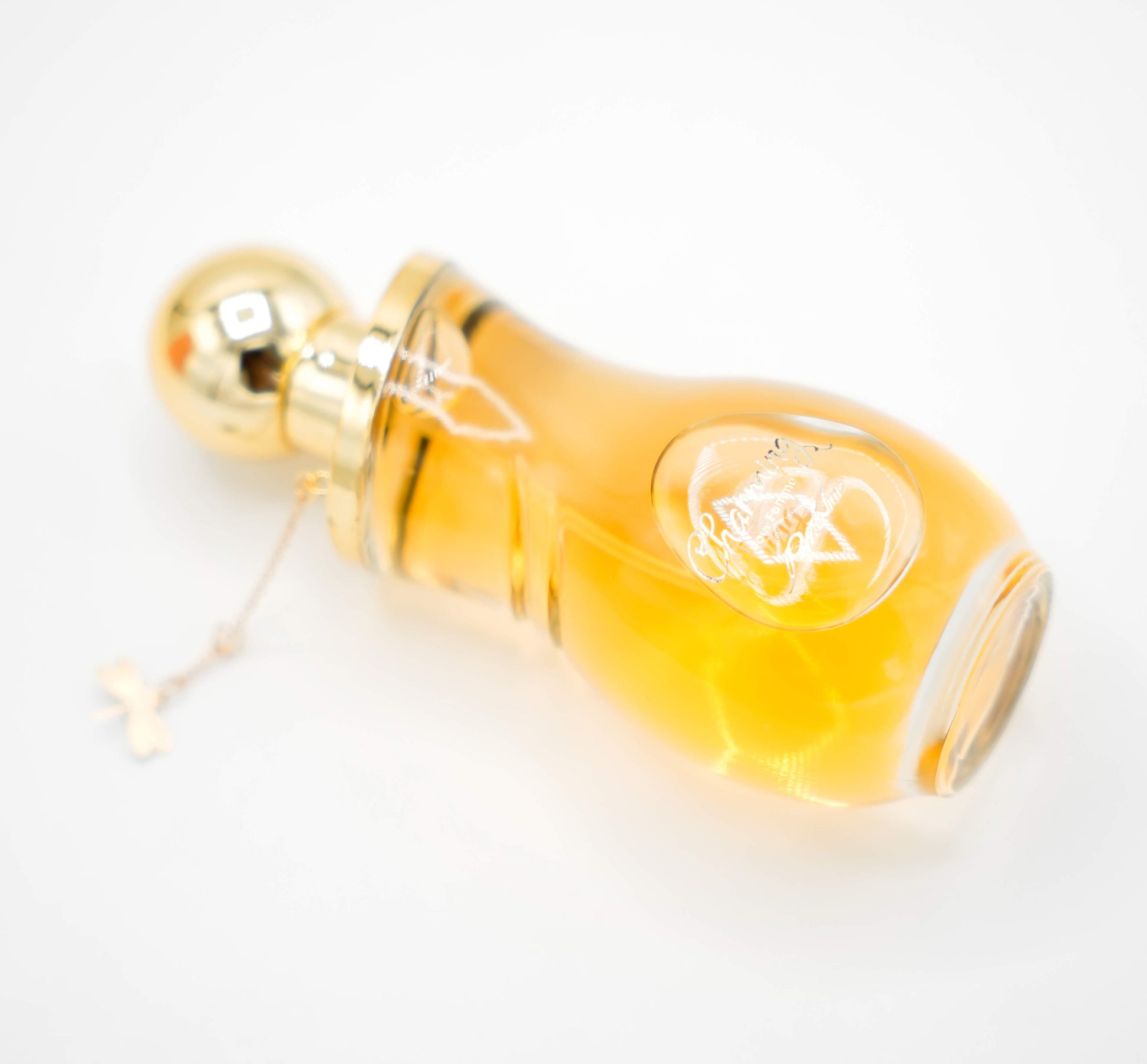 A 100ml bottle of 'CHARMING EDP' for women displayed on a white pedestal with a yellow liquid inside. The bottle has a golden cap and there is a green leaf to the top right of the image.
