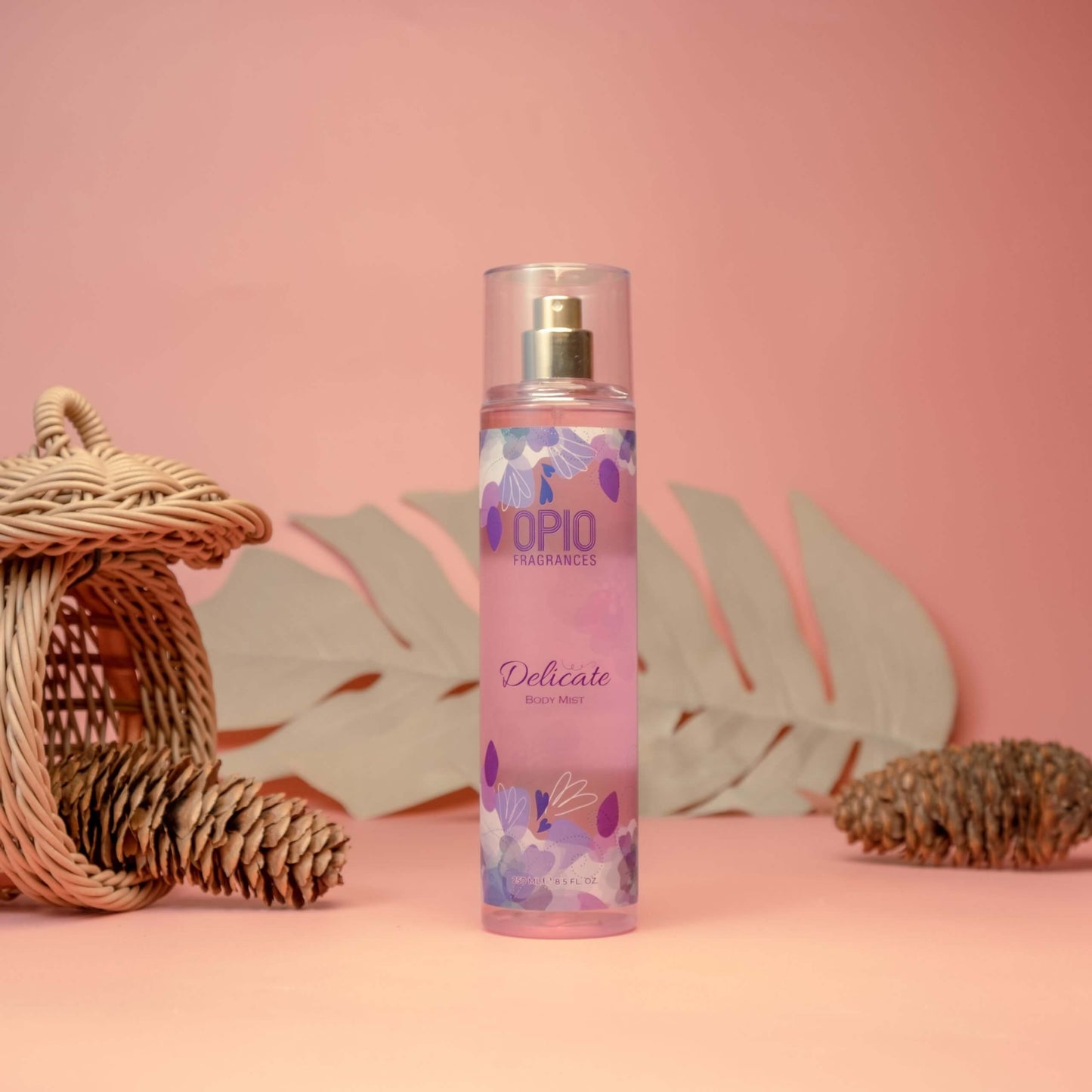 A bottle of DELICATE BODYMIST for women, with a pink label and floral design, placed on a surface next to a wicker basket and a flower.
