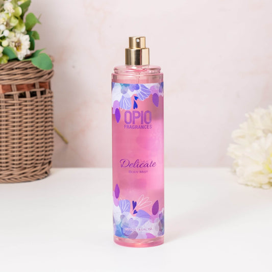A bottle of DELICATE BODYMIST for women, with a pink label and floral design, placed on a surface next to a wicker basket and a flower.