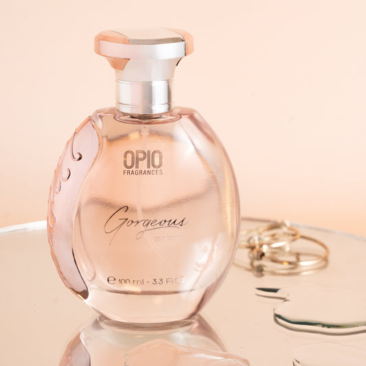 A bottle of OPIO Gorgeous women's fragrance, with a clear, pink-colored liquid visible inside. The bottle is placed on a reflective surface, and a set of keys is partially visible in the foreground.