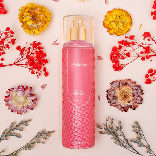 A pink bottle of body mist for women surrounded by various flowers and foliage on a white background.