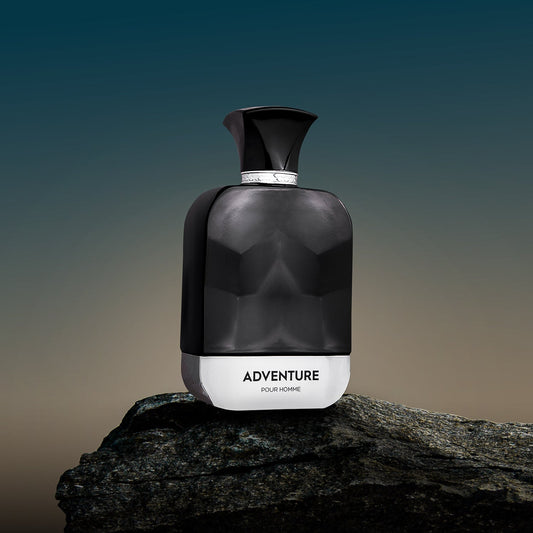 A spray bottle of 'ADVENTURE FOR MEN' perfume, with a dark, smoky design, placed on a rock against a sunset background.