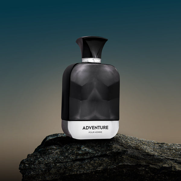 A spray bottle of 'ADVENTURE FOR MEN' perfume, with a dark, smoky design, placed on a rock against a sunset background.