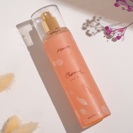 A pink bottle of Perrie Vian by OPIO Charming Body Mist for Women, with a clear liquid visible inside the bottle and a gold-colored cap. The bottle is placed on a white surface with some feathers around it, and flowers are also present in the background.