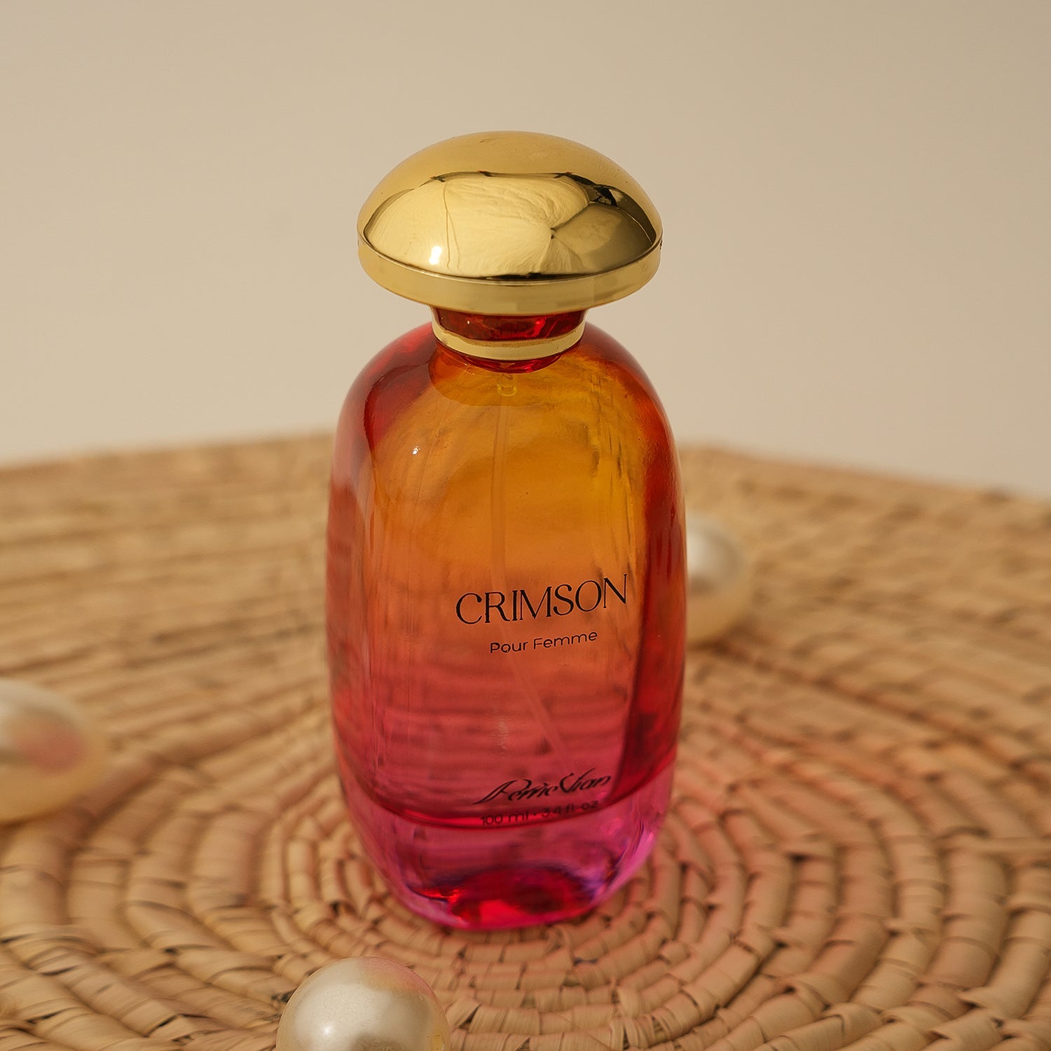 A glass bottle of CRIMSON EDP for women with a gold-colored cap, placed on a woven surface. The bottle has a gradient of orange to pink color.