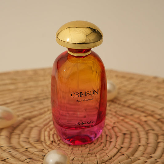 A glass bottle of CRIMSON EDP for women with a gold-colored cap, placed on a woven surface. The bottle has a gradient of orange to pink color.
