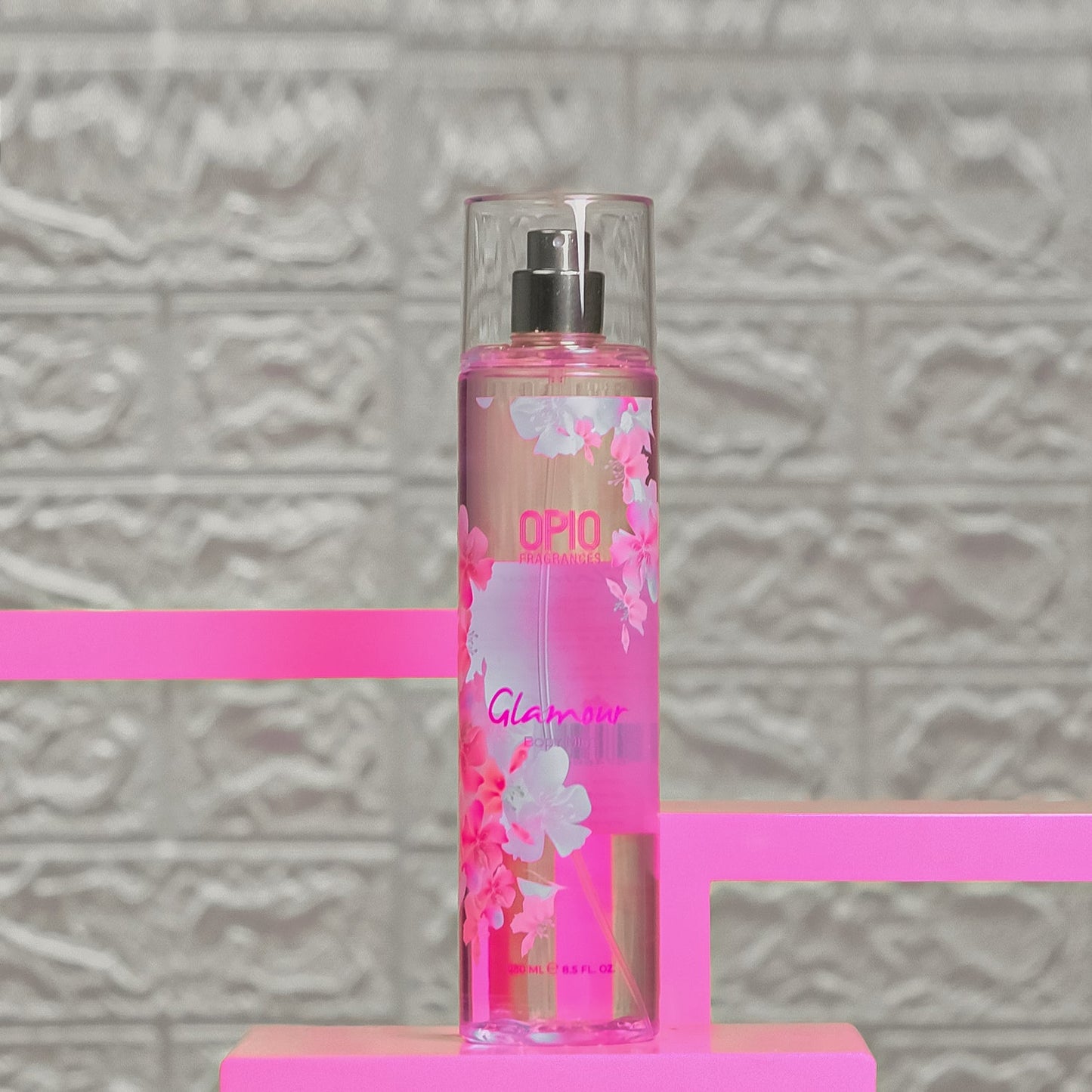 A bottle of 'GLAMOUR' body mist for women with a floral design, placed on a pink surface with a patterned background.