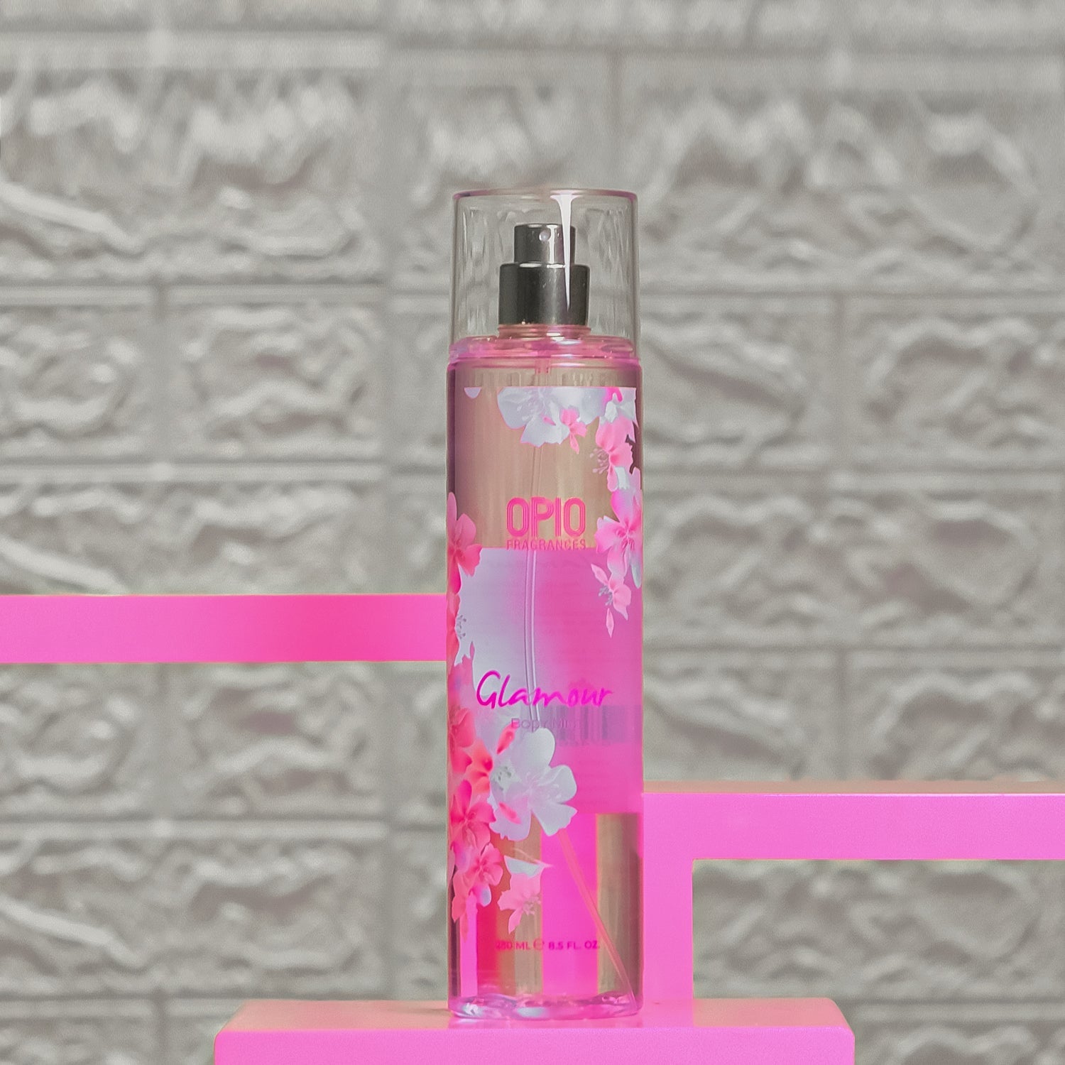 A bottle of 'GLAMOUR' body mist for women with a floral design, placed on a pink surface with a patterned background.